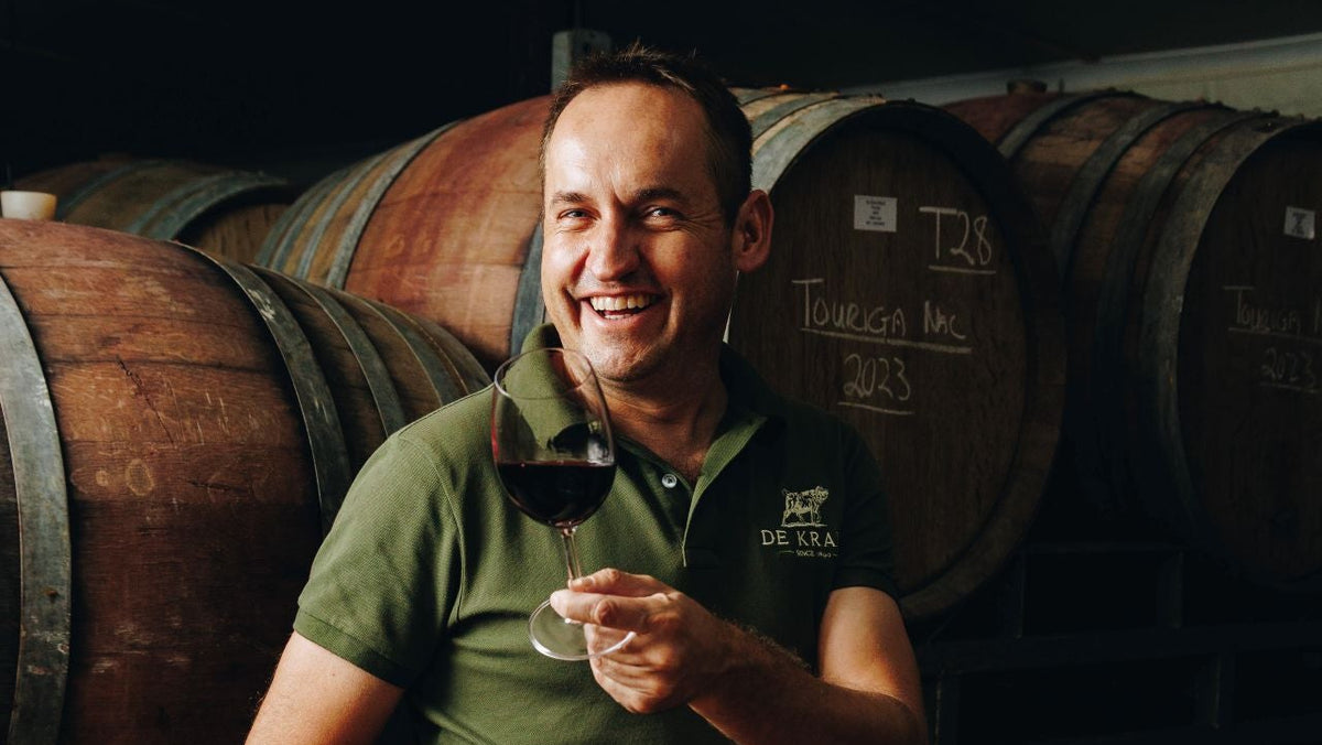 A smiling man in a green polo shirt and khaki shorts sits on a wooden wine barrel in a winery, holding a glass of red wine. More wooden barrels are stacked in the background.